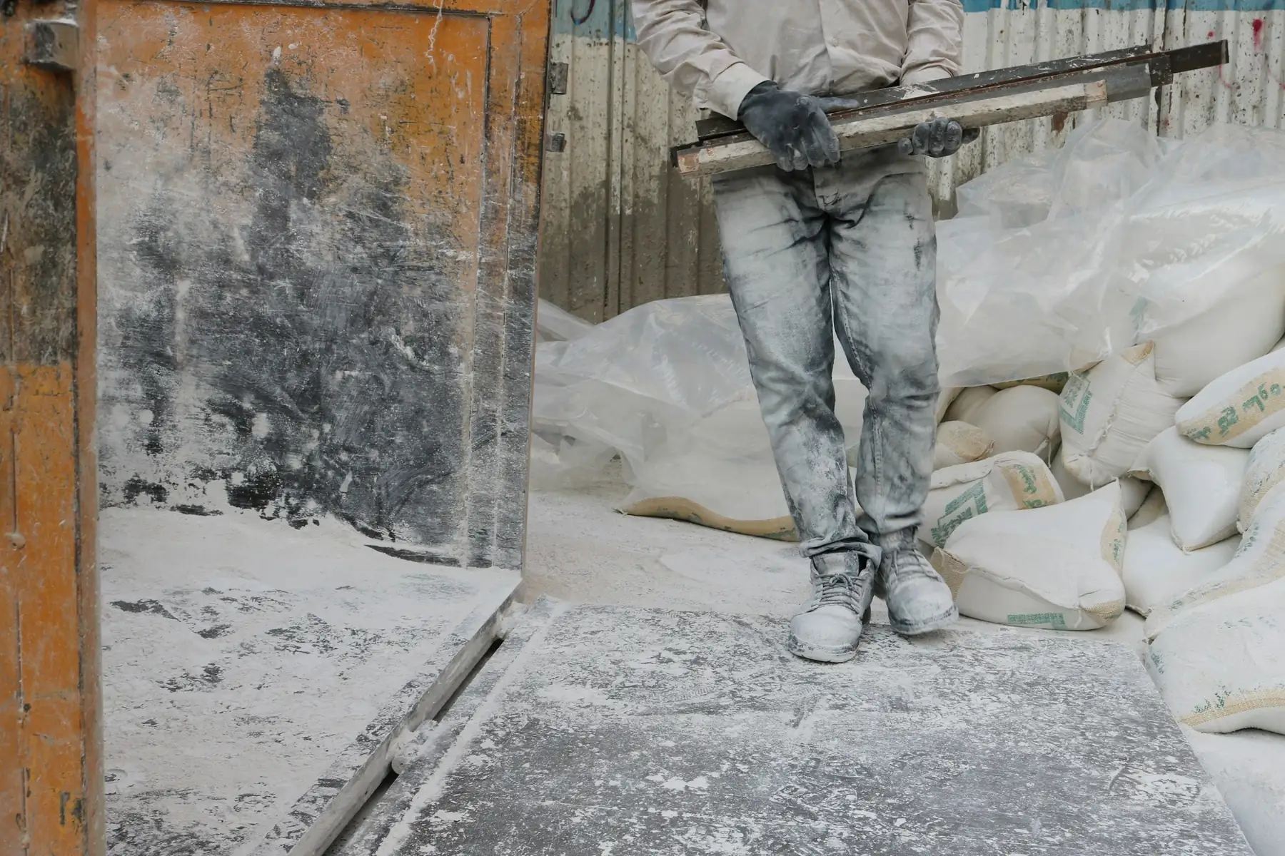Opérateur en combinaison qui projette un abrasif, sacs de sable et poussière blanche autour